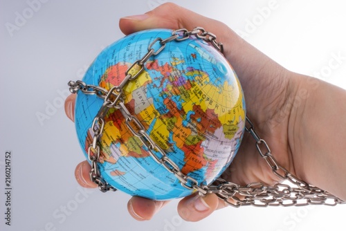 Foto Human hand holding small terrestrial globe secured by metal chain against neutra