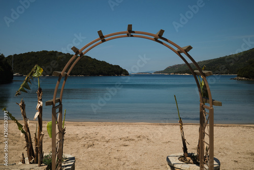 Wooden beach arch overlooking a tranquil Mediterranean bay, beautiful Sivota bay landscape in Greece, scenic Ionian sea coast with turquoise water and green island, Mediterranean summer vacation