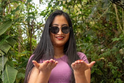 Woman smiling, wearing sunglasses, experiencing mindo's rich biodiversity, feeding a hummingbird directly from her open hand in a tropical rainforest setting