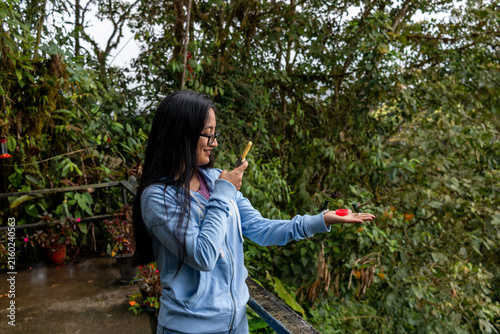Young woman enjoying a close up interaction with a group of hummingbirds, holding out her hand to feed them while capturing the moment on a smartphone in the lush mindo cloud forest