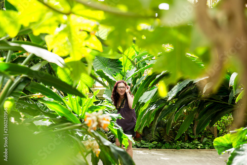 Cheerful Woman Touching Eyeglasses and Smiling in a Lush Tropical Greenhouse surrounded by Dense Foliage