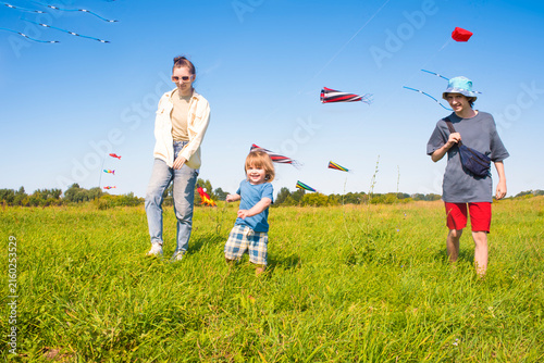  selective focus on a happy toddler boy walking with his teenage siblings on a green field with colorful kites flying in the blue sky behind them on a summer day.