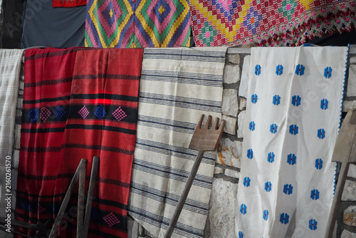 Fotografía Display of vibrant, traditional woven Greek textiles, including red and white blankets and rugs with geometric patterns, hanging on a stone wall next to rustic wooden farming tools