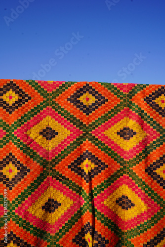 Fotografie Vertical shot of vibrant handwoven kilim rug with colorful diamond patterns hanging outdoors under a clear blue sky