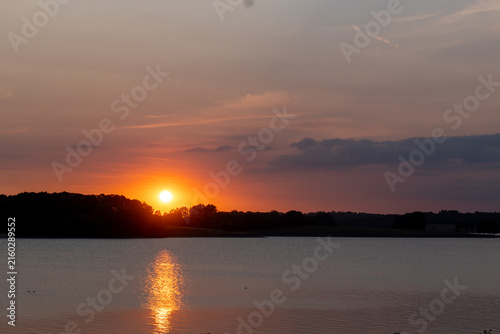 Obraz na plátne Orange sun setting over calm water with a golden reflection and silhouetted shoreline