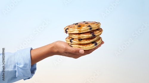 Person holding stack of fluffy blueberry pancakes drizzled with sweet syrup against soft blue background with stack four warm