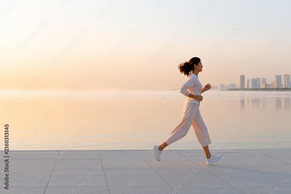 Fototapeta premium Woman running along a calm seaside promenade at sunrise with city skyline in the distance for wellness background and active lifestyle banner design