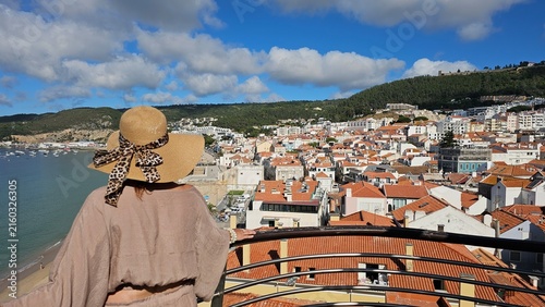 Elegant woman in straw hat with leopard bow and pearl bracelets looking at sunny beach in Portugal