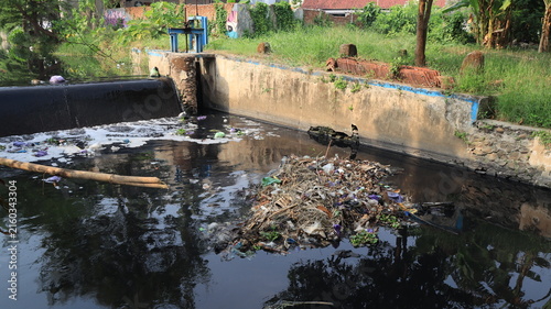 Foto A residential canal carries visibly polluted dark water and floating plastic, bordered by concrete walls, trees, and houses in a densely developed neighborhood