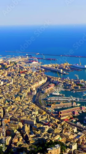 Vertical video. Genoa, Italy. Aerial panorama over Porto Antico, where historic rooftops meet the deep blue Ligurian Sea under bright summer light, with the white panoramic wheel marking the revitali