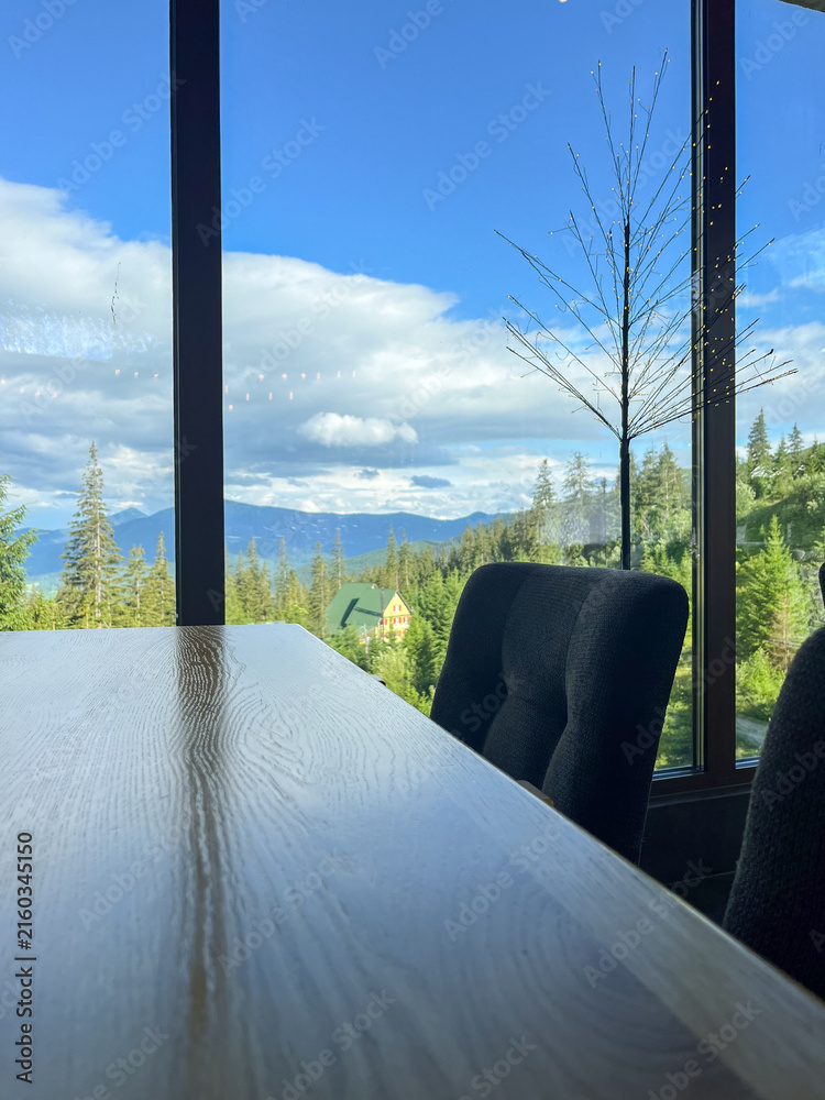 Fototapeta premium Cozy interior with panoramic view of mountain forest and blue sky, a wooden table in foreground