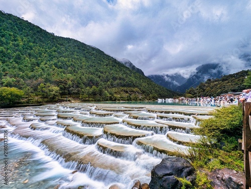 View of the turquoise terraced waterfalls of Blue Moon Valley surrounded by lush green mountains and a cloudy sky in Lijiang, Yunnan, China.