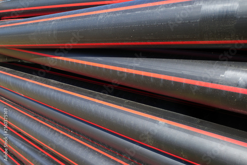 Stacked high-density polyethylene (HDPE) pipes are shown, typically used for cable protection. The pipes are black with distinctive red stripes running along their length. 