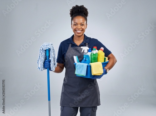 Smiling young woman cleaner holding a mop and a basket full of colorful cleaning supplies and chemical bottles for professional housekeeping services