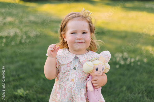 little 2‑year‑old girl is on the lawn in summer at sunset, holding a princess doll.