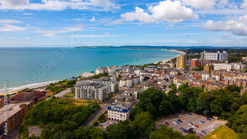 Aerial view of the Bournemouth coastline featuring the sandy beach, urban architecture, and lush greenery under a bright blue sky, Bournemouth, England, United Kingdom.