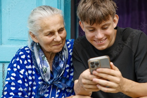Принти на полотні Happy teenage grandson and senior grandmother looking at mobile phone screen, smiling and sharing warm family moment outside