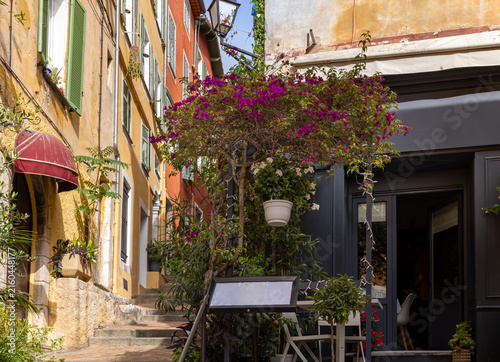 View of the narrow streets of Villefranche Old City, featuring colorful buildings, purple bougainvillea, and a restaurant, Villefranche-sur-Mer, Provence-Alpes-Cote d'Azur, France.