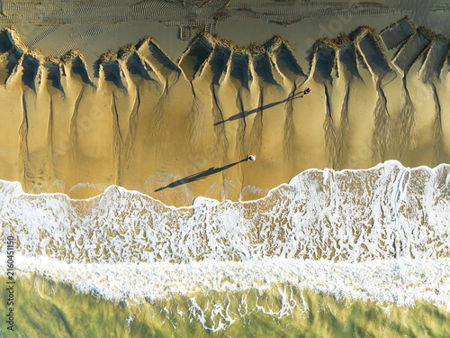 Aerial view of Ellis Beach with unique geometric patterns in the sand, crashing waves, and two people casting long shadows on the shoreline in Cairns, Queensland, Australia.