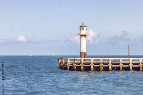 A serene coastal scene features a solitary lighthouse perched on a weathered pier, extending into the calm, the sea under a bright sky, with distant sailboats. Nieuwpoort town in Belgium, Europe.