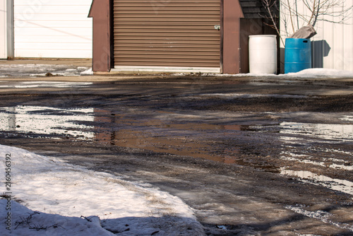 The Muddy Moat: When Your Storage Unit Comes with a Complimentary Water Feature. A wide shot of a commercial storage facility or industrial outbuilding driveway during a messy spring thaw.