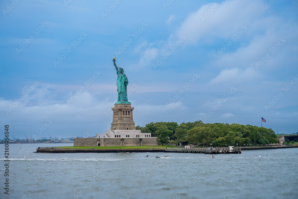 Obraz premium Statue of Liberty on Liberty Island viewed from Hudson River. Copper statue on stone pedestal surrounded by green trees under cloudy sky with rippling water in foreground.