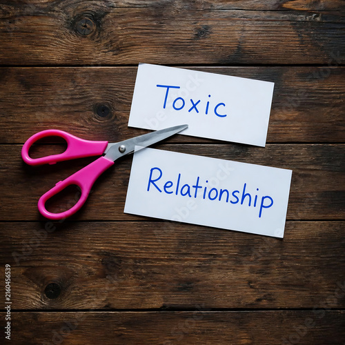 Scissors cutting toxic relationship label in half. Conceptual breakup symbol with handwritten note on wooden background.