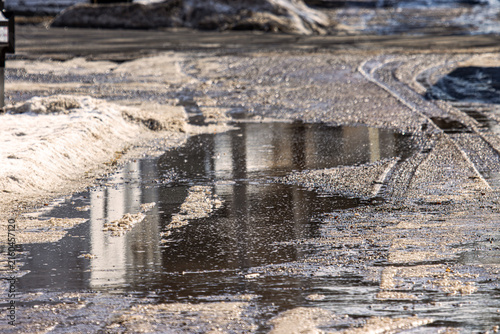 The Muddy Crossroads: When Spring Thaw Decides to Make Its Own Route. A wide shot of a rugged, wet gravel road or boat launch area during a spring thaw, featuring a large, murky puddle.
