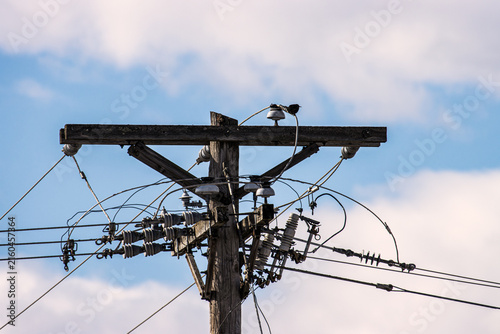 A close-up view of the top of a weathered wooden utility pole, showcasing a complex network of crossarms, porcelain electrical insulators, transformers, and a messy tangle of overhead power lines.