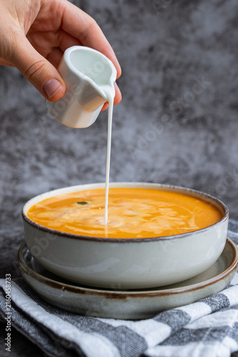 Smooth texture natural colors and fresh dairy contrast beautifully with the garnish of Creamy Pumpkin Soup Garnished with Pumpkin Seeds Cream Pouring by Hand. Concept of homemade cooking seasonal