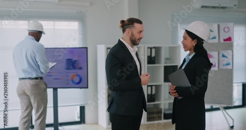 Portrait of bearded manager discusses infrastructure development with female engineer in office. Colleagues talk while construction inspector works in boardroom