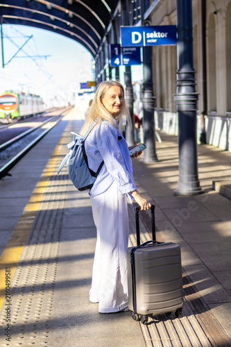 Mature woman walks toward train platform holding smartphone while pulling rolling suitcase behind her. Concept of solo business travel and digital navigation during commute to train departure.