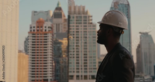 African American architect wearing a white hardhat stands in side profile against a modern skyline, representing construction expertise and urban development.