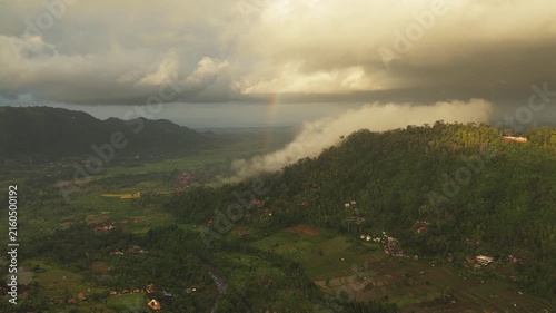Aerial view of Sidemen Rice Terrace with lush green hills, misty clouds, and a faint rainbow over the valley in Kecamatan Sidemen, Bali, Indonesia.