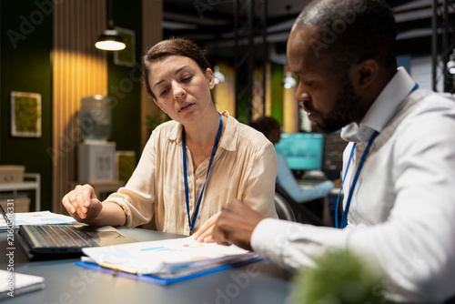 Фотография Manager supervises an employee in the office and evaluates staff performance and task completion on a laptop, comparing metrics on a notebook while overseeing the businesswoman's work at night