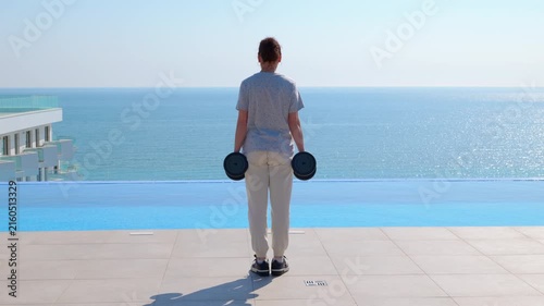 Woman walking with dumbbells near infinity pool and sea. Woman performing a farmer's walk exercise with dumbbells along the edge of an infinity pool, with a sea view in the background on a bright
