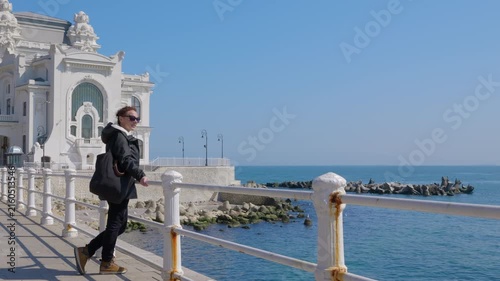 Female tourist walking on seaside promenade admiring the constanta casino. Young woman wearing a backpack walking on a seaside boardwalk on a sunny day. Stopping to admire the view of the black sea