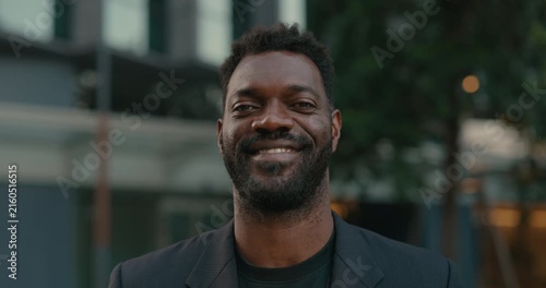 Smiling African American businessman looks at the camera outside a contemporary office, conveying approachable leadership, confidence and professional success.
