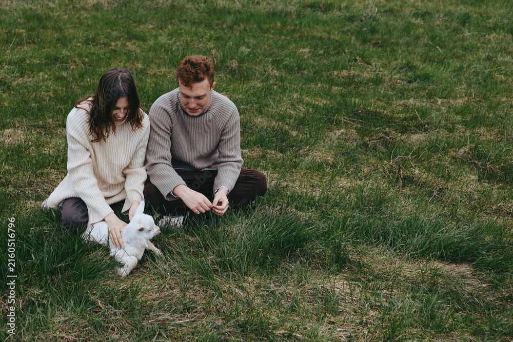 Fototapeta premium A couple sits on a grassy meadow with a goat kid, enjoying a calm outdoor moment, a wholesome lifestyle scene that highlights family, nature, companionship, and simple rural joy.