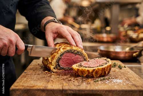 Chef hands slicing perfectly baked hot Beef Wellington with steam on wooden board in professional kitchen