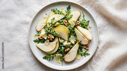 A fresh gourmet salad featuring sliced green pears, crisp arugula leaves, crunchy walnuts, and parmesan cheese shavings served on a white ceramic plate background.