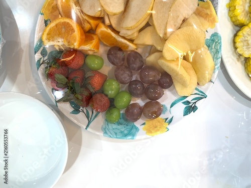 Fotografía Colorful assorted fresh fruit platter covered in clear plastic cling wrap, arranged on floral ceramic plate alongside white bowl