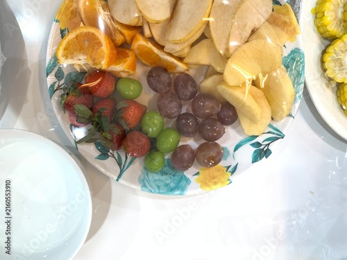Obraz na płótnie Colorful assorted fresh fruit platter covered in clear plastic cling wrap, arranged on floral ceramic plate alongside white bowl
