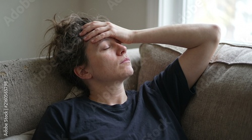 A woman resting on a couch with her eyes closed and hand on her forehead, appearing stressed or unwell. The soft lighting suggests a quiet, possibly indoor setting.