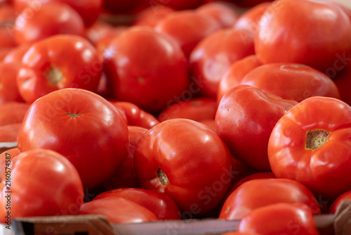 A harvest of large, fresh organic tomatoes stockpiled in a crop bunch for sale at a farmers' market. The raw tomatoes are stacked, exposing the bright red color of the vegetables with smooth, shiny sk