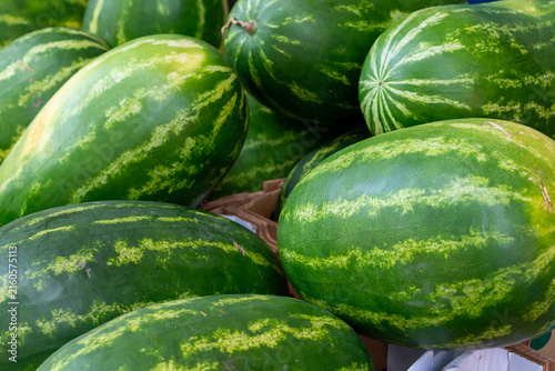 A bulk supply of large, organic, oblong shaped green and yellow striped watermelons on a table at a farmers' market. The African melons have thick peels, with a striped pattern and a juicy interior. 