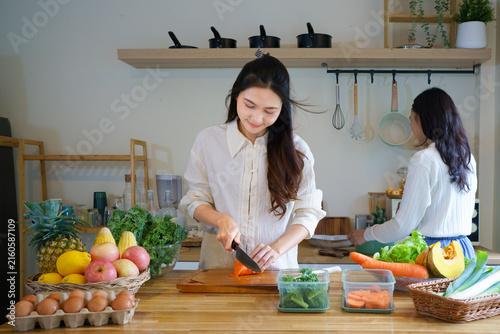 Two smiling Asian women cheerfully chop and prep colorful fresh vegetables and fruits in a bright home kitchen, ideal for healthy lifestyle and nutrition content