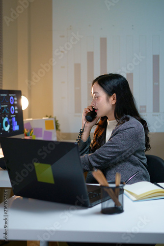 Asian Businesswoman Talking on Phone at Office Desk with Laptop and Data Dashboard at Night, business communication and corporate work concepts.