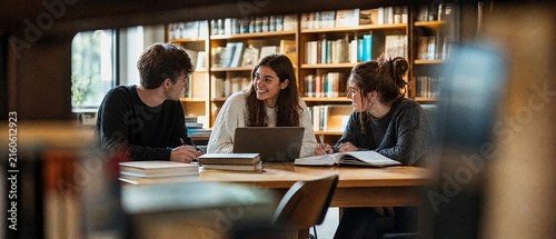 College student study group collaborating around laptop and books in warm library, sharing ideas with focused expressions and teamwork during academic learning session