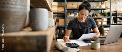 Ceramic artisan reviewing paperwork in pottery studio with laptop and calculator